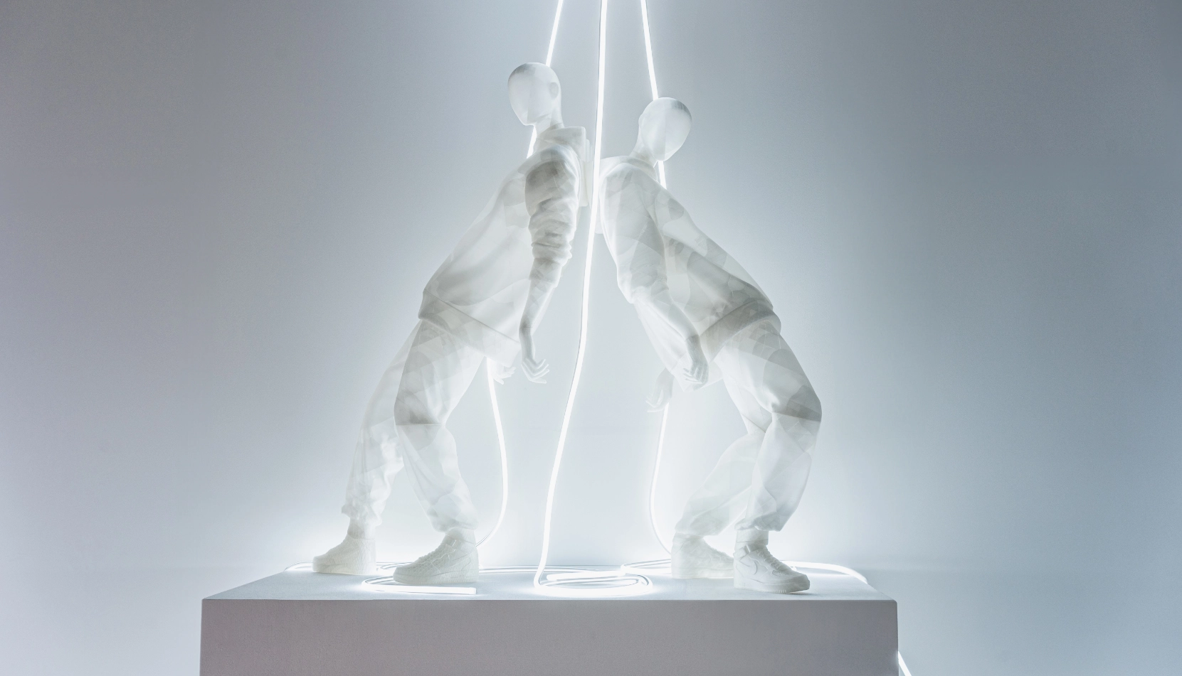 Two translucent white mannequins from the Hans Boodt Blend — Frozen in Time collection face each other in a wide, grounded stance on a white plinth. A glowing neon light strip runs vertically between them and descends in a loop at their feet, illuminating their semi-transparent forms against a soft grey background.