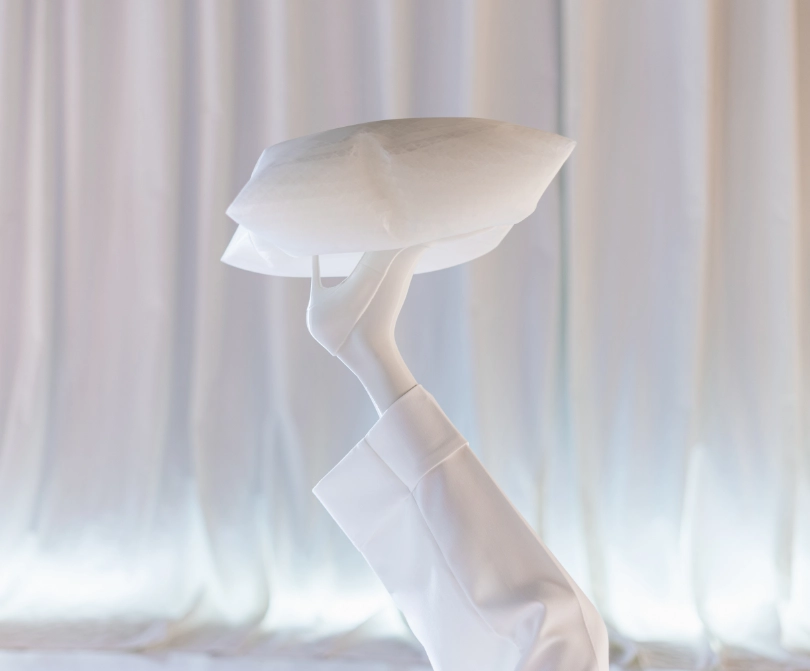 Close-up of a white mannequin arm and hand from the Blend — Frozen in Time collection, raised to balance a geometric faceted head form on the fingertips. Shot against softly draped white fabric.