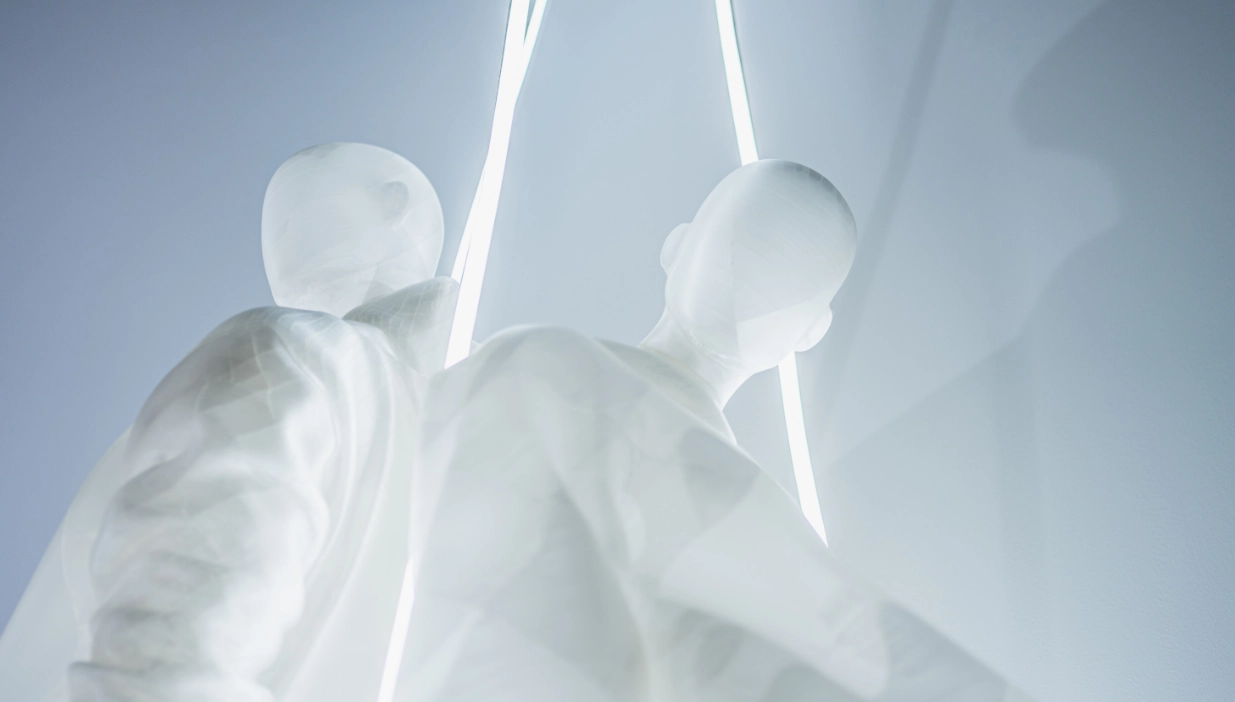 Low-angle close-up looking up at two translucent white mannequins from the Blend — Frozen in Time collection, their backs turned toward each other as a glowing neon strip rises between them into the light blue background. The shot emphasises the ethereal, ghostly quality of the semi-transparent material.