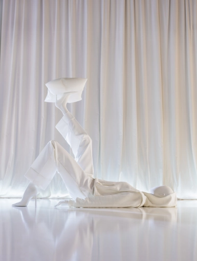 A white mannequin from the Blend — Frozen in Time collection reclines on a reflective white floor, one leg raised and bent, balancing a geometric faceted head form on its foot. The pose is dynamic and sculptural, set against a backdrop of white draped fabric.