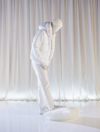 A white male mannequin from the Blend — Frozen in Time collection dressed in an all-white puffer vest, hoodie, and trousers stands in a deep forward lean, head bowed. Shot against floor-to-ceiling white curtains on a reflective white floor.