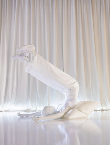 A white mannequin from the Blend — Frozen in Time collection in a crouched, arched pose with one leg extended and a sneaker raised into the air. The body twists dramatically toward the floor, with a geometric faceted head resting beside it. Shot against white draped fabric on a reflective surface.