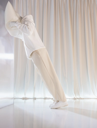 A white mannequin from the Blend — Frozen in Time collection in a strong forward lean, arms folded across the torso and head tucked down, dressed in white wide-leg trousers and sneakers. The bottom half of the figure is in focus; the upper body tapers to a sharp angular point. Shot against white curtains on a reflective floor.