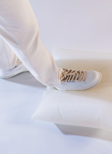 Close-up of a white mannequin foot and lower leg from the Blend — Frozen in Time collection, wearing a cream sneaker with beige laces, stepping onto a soft white form on a light grey surface.